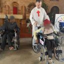 padre con tonaca bianca e croce rossa che spinge donna in sedia a rotelle. Uomo su sedia a rotelle sullo sfondo