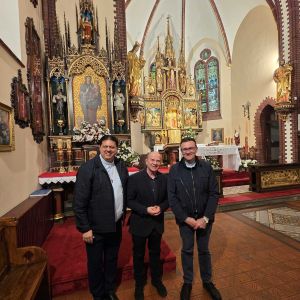 Tre uomini vestiti di nero posano davanti aun altare in oro con l'immagine della madonna. Uno degli uomini ha al collo una catenina con la croce rossa dei camilliani