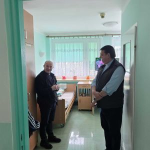 Padre Pedro Tramontin, superiore generale dei Camilliani, visita con un uomo vestito di nero la stanza di un ospedale. Le pareti sono verdi chiaro
