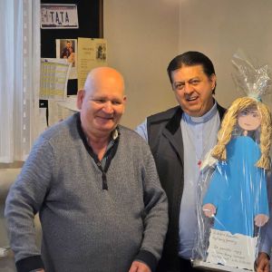 Padre Pedro Tramonti nel suo viaggio in Polonioa riceve in dopno una bambola azzurra con i capellio biondi. Padre Pedro ha una camicia grigia, un gilet nero e una collanina con una croce rossa. Accanto a lui c'è un uomo pelato con un maglione grigio