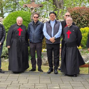 sette uomini posano per una foto in un aprco. due hanno gli occhiali da sole. tre sono vestiti da preti con un abito nero e una croce rossa sul petto tipica dei camilliani