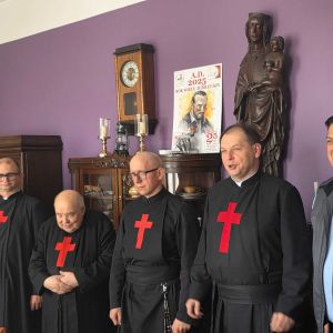 Foto di 6 uomini davanti a una stata di legno della madonna. Quattro uomini indossano l'abito nero dei camilliani con la croce rossa sul petto. Uno ha una catenina con la croce rossa dei camilliani. Le pareti sullo sfondo sono viola