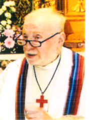 Uomo anziano con abito bianco, stola a strisce di diversi colori e croce rossa sul petto. L'uomo ha gli occhiali e i capelli bianchi.