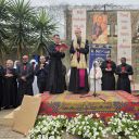 Alcuni regliosi vestiti di nero con una croce rossa sul petto sono intorno a un cardinale che parla sul palco. Il cardinale ha un berretto fucsia in testa. Il palco è rivestito di tappeti e in basso ci sono dei fiori.