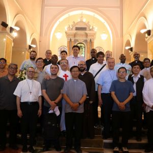 Un gruppo di uomini è disposto su tre gradini di un altare. Molti hanno una croce rossa sul petto. Qualcuno è vestito da sacerdote. Ai lati e sul retro ci sono delle arcate illuminate