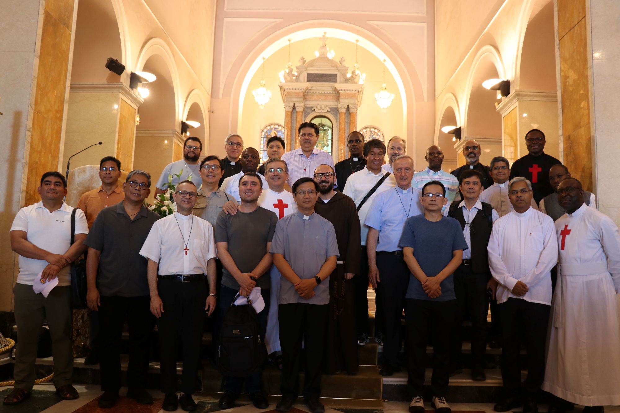 Un gruppo di uomini è disposto su tre gradini di un altare. Molti hanno una croce rossa sul petto. Qualcuno è vestito da sacerdote. Ai lati e sul retro ci sono delle arcate illuminate