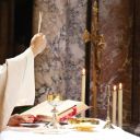 Un sacerdote con una tunica bianca alza un'ostia in alto con una mano. L'altra mano è sopra un piattino d'oro. Davanti a lui c'è un calice d'oro, un libro e alcune candele. Sullo sfondo ci sono delle colonne di marmo grigio e marroni