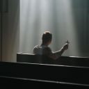 Un uomo è seduto in chiesa e ha in mano una piccola croce. Un fascio di luce lo illumina dall'alto. Dietro di lui ci sono i banchi per sedersi