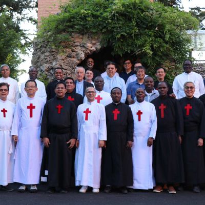 Gruppo di religiosi camilliani. Alcuni hanno la tunica sacerdotale bianca, altri hanno la tunica nera. Tutti hanno la croce rossa di san Camillo sul petto. Sullo sfondo ci sono degli alberi.