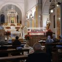 In una chiesa, dal pulpito, un uomo con una camicia bianca parla alle persone sedute tra i banchi. Sull'altro lato della chiesa c'è una bara in vetro con il corpo di san Camillo vestito di nero e rosso come i camilliani