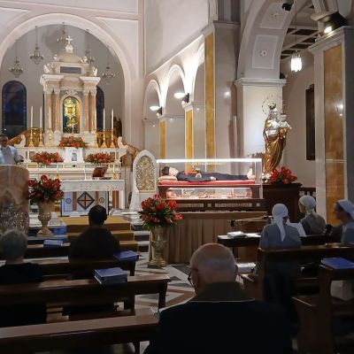 In una chiesa, dal pulpito, un uomo con una camicia bianca parla alle persone sedute tra i banchi. Sull'altro lato della chiesa c'è una bara in vetro con il corpo di san Camillo vestito di nero e rosso come i camilliani