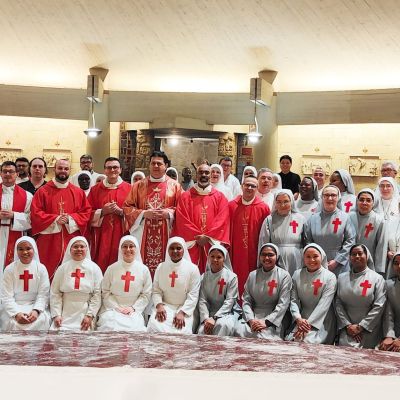 Un gruppo di suore e di preti sono davanti all'altare di una chiesa per una foto. La suore sono vestite di bianco o di grigio e hanno un a croce rossa sul petto. Alcuni preti hanno una stola rossa, altri hanno un abito rosso da cerimonia