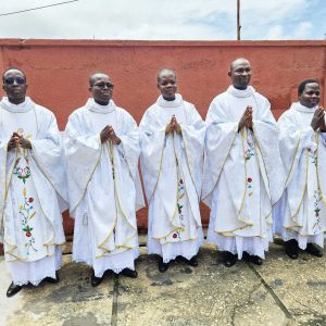 Cinque uomini africani posano con le mani giunte davanti a un muro marrone. sopra di loro il cielo è nuvoloso. gli uomini sono vestiti di bianco con l'abito dei sacerdoti. i vestiti sono tutti bordati d'oro e hanno dei fiori disegnati al centro