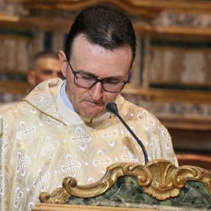 Un uomo vestito di bianco con gli occhiali parla in chiesa davanti a un microfono