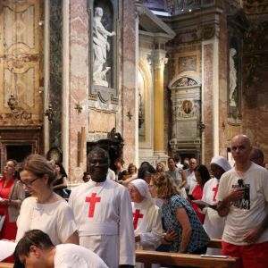 Un gruppo di persone ascolta la messa. Alcuni hanno un vestito bianco da preti o da suore e una croce rossa sul petto