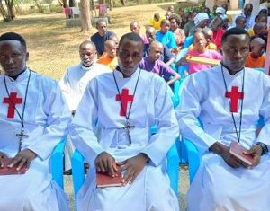 Tre uomini africani vestiti di bianco con la tunica dei preti e la croce rossa di san Camillo sono seduti su delle sedie azzurre. Hanno tutti in mano un libretto marrone