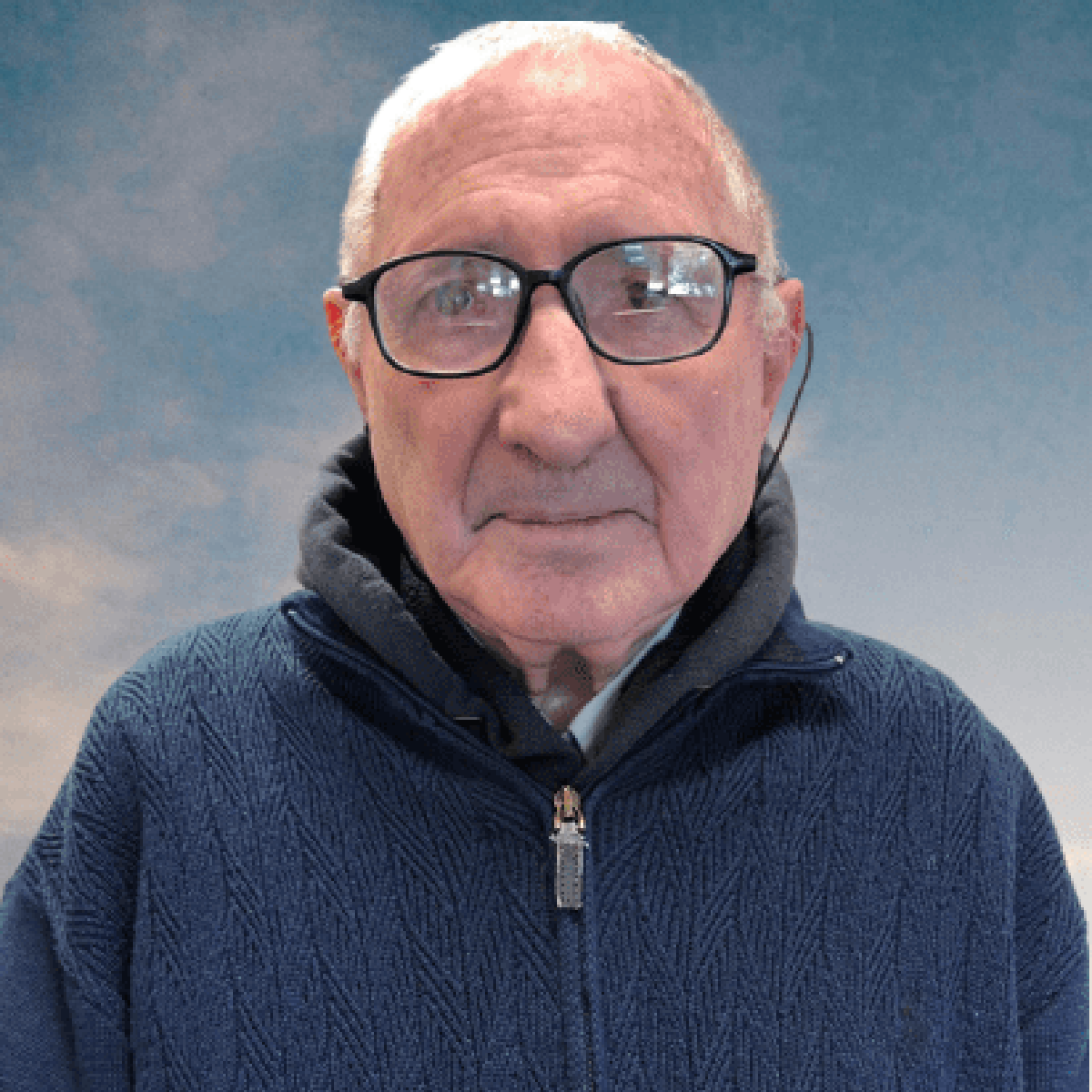 Primo piaNo di un uomo anziano con gli occhiali bordati di nero. Indossa una giacca blu. Lo sfondO è un cielo nuvoloso