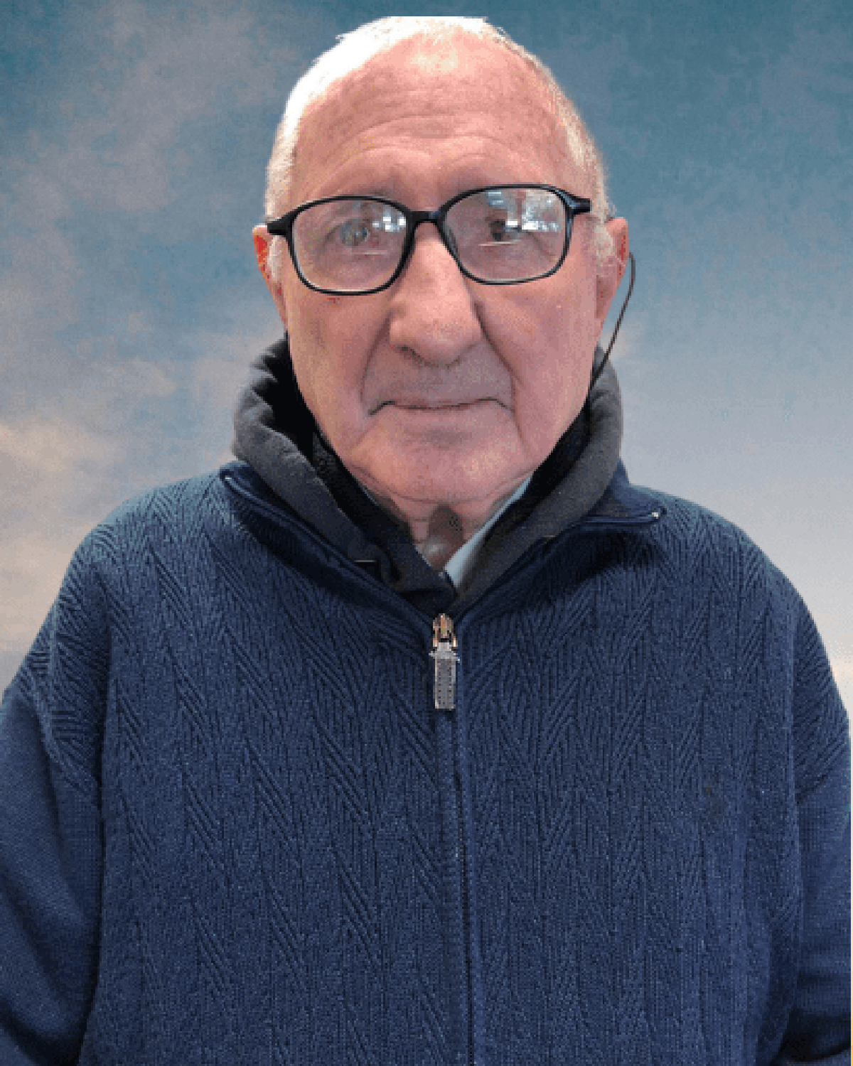 Primo piaNo di un uomo anziano con gli occhiali bordati di nero. Indossa una giacca blu. Lo sfondO è un cielo nuvoloso