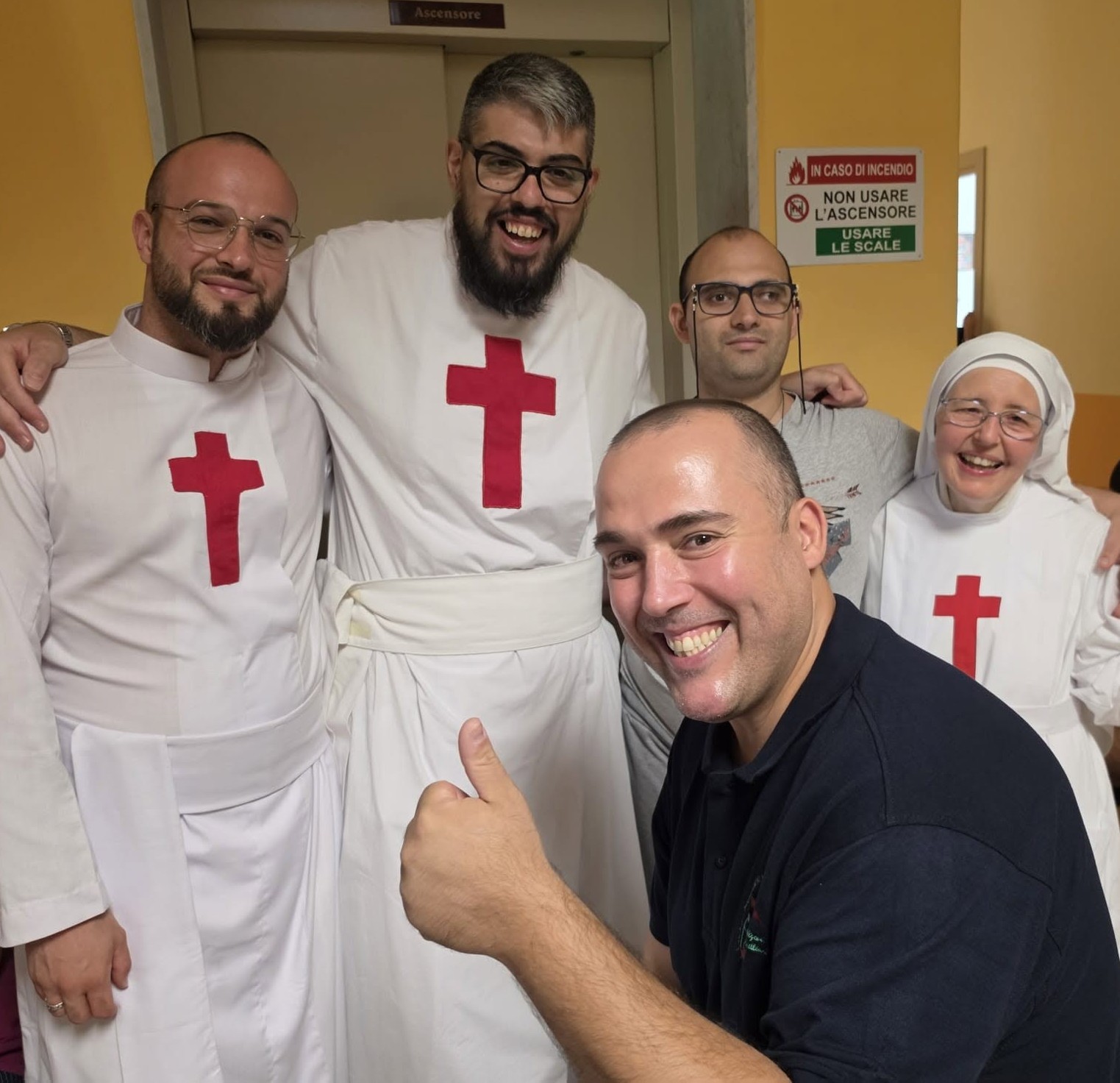 Nella foto ci sono cinque persone. Tre persone - due omini e una donna- sono dei religiosi camilliani con l'abito bianco e la croce rossa sul petto. In prima fila c'è un uomo vestitomdi nero che sorride. Sullo sfondo emerge la testa di una quinta peersona. Dietro a tutti le pareti sono gialle. La foto è scattata all'interno di una stanza.