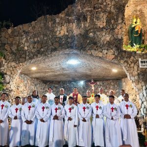 Un gruppo di uomini è disposto su due file. Davanti ci sono uomini vestiti di bianco con la croce di san Camillo e una coccarda di fiori sul eptto. Dietro ci sono sacerdoti con tuniche gialle o bianche. Sullo sfondo ci sono delle rocce, un crocefisso e una statua della Madonna con bambino.