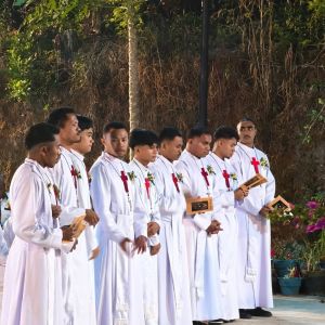 Nove uomini sono vestiti di bianco con la tunica da preti e hanno una croce rossa sul petto. Hanno tutti una spilla a forma di fiore, un libretto marrone per le letture e una catenina con una croce d'argento. Sullo sfondo ci sono degli alberi e dei fiori