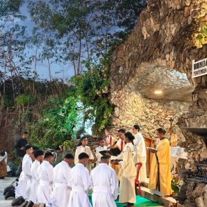 Alcuni giovani vestiti di bianco sono incinocchiati davanti a un sacerdote vestito di bianco. Sulle maniche ha delle strisce rosse. Dietro a lui ci sono due altri sacerdoti. Ai lati ci sono tre ragazze chierichetto. Sullo sfondo ci sono delle rocce e degli alberi. il cielo è azzurro scuro e nuvoloso.