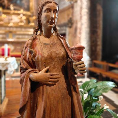 Nell'immagine c'è la statua in legno di una donna con i capelli sciolti, un calice in mano e una mano sulla pancia. Ha un vestito lungo drappeggiato sulle spalle e sulle braccia. Dietro di lei ci sono delle piante che le arrivano al fianco e un altare