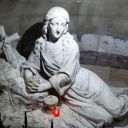 Una statua raffigura una donna semisdraiata su dei sassi. Vicino alla sua testa c'è il crocefisso. Vicino le mani allacciate c'è una lampada votiva rossa accesa. Tutta l'opera è sui toni del grigio.