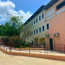 Nela foto si vede un palazzo in psospettiva con una parte giallo chiara e una parte arancione. Ci sono due file di fin estre contornate di bianco. Sullo sfondo c'è il cielo nuovoloso e degli alberi. Sul davanti c'è una strada in terra gialla e una ringhiera in ferro.