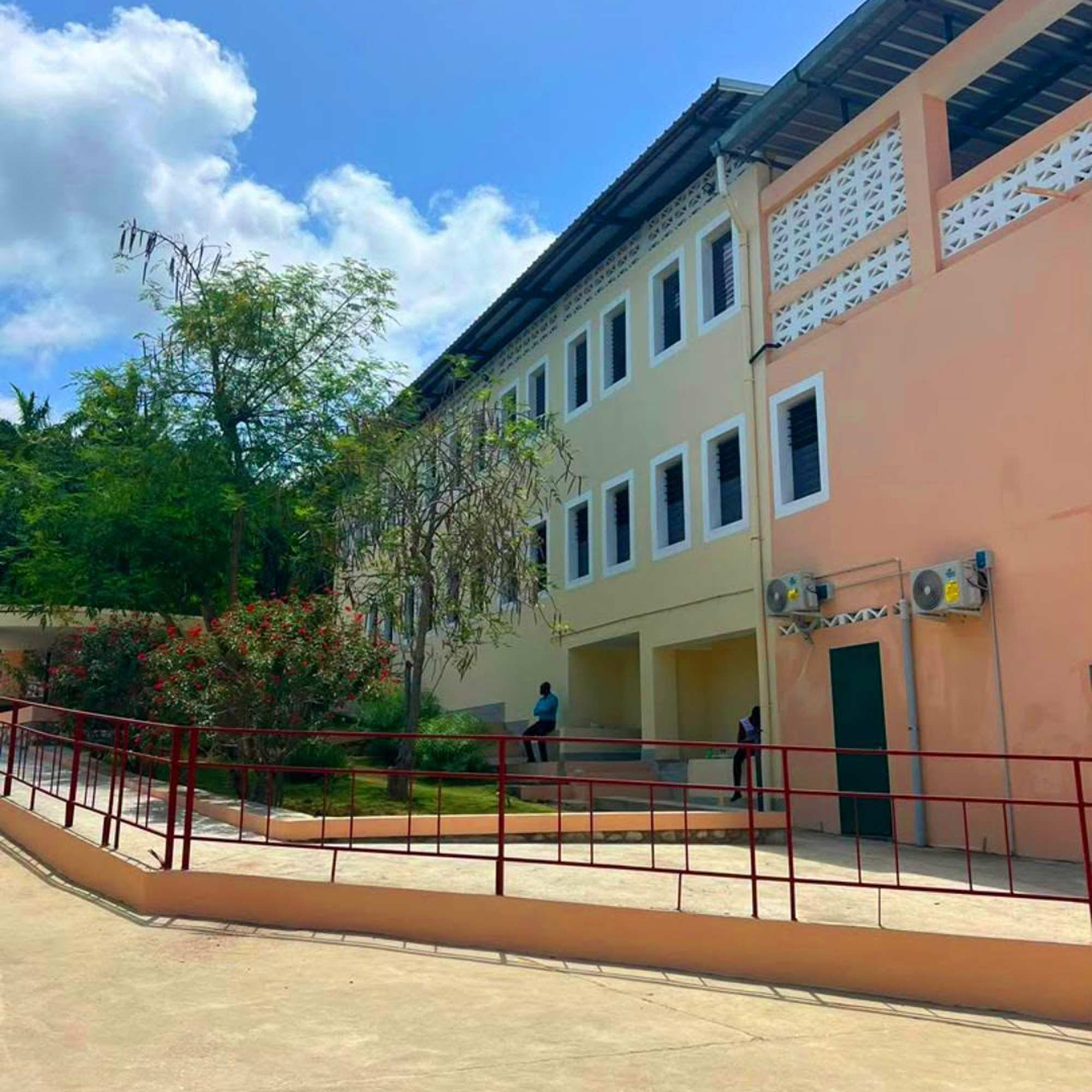Nela foto si vede un palazzo in psospettiva con una parte giallo chiara e una parte arancione. Ci sono due file di fin estre contornate di bianco. Sullo sfondo c'è il cielo nuovoloso e degli alberi. Sul davanti c'è una strada in terra gialla e una ringhiera in ferro.