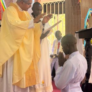 Un uomo africano è inginocchiato davanti a un sacerdote vestito di giallo. Accanto all'uomo vestito di giallo c'è un altro sacerdote vestito di bianco. Sullo sfondo, un uomo seduto vestito di bianco con la croce rossam legge un testo. A terra c'è un tappeto rosso.