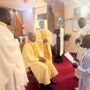 Un uomo africano è inginocchiato davanti a un sacerdote seduto vestito di giallo. Accanto all'uomo vestito di giallo ci sono altri due sacerdoti vestiti di giallo. Accanto all'uomo in ginocchio c'è un uomo vestito di bianco con la stola con la scritta Ministri degli infermi - san Camillo e una croce rossa.