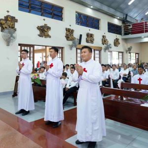 Tre giovani uomini asiatici pregano in una chiesa in piedi. Dietro di loro ci sono altri giovani seduti nei banchi della chiesa. Alcuni hanno l'abito di san Camillo con la croce rossa. In alto c'è un coro di donne vestite con abiti rosa intenso