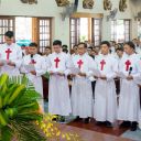 Un gruppo di giovani uomini vestiti con la tunica bianca dei sacerdoti legge un foglio davanti a un altare di una chiesa. Hanno tutti i capelli neri. Il loro abito hauna croce rossa di san Camillo sul petto. Sul lato ci sono delle finestre aperte. Davanti a loro c'è una pianta. Il pavimento è di legno marrone lucido