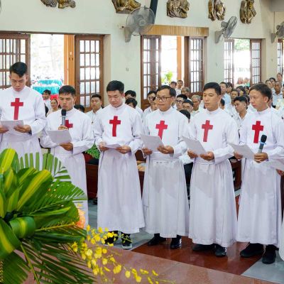 Un gruppo di giovani uomini vestiti con la tunica bianca dei sacerdoti legge un foglio davanti a un altare di una chiesa. Hanno tutti i capelli neri. Il loro abito hauna croce rossa di san Camillo sul petto. Sul lato ci sono delle finestre aperte. Davanti a loro c'è una pianta. Il pavimento è di legno marrone lucido