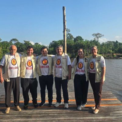 Sei persone sono in posa per uanfoto su un molo di elgno arancione davanti al fiume. Hanno una maglietta bianca con la scritta Camilianos e portano pantaloni neri. Nella foto ci sonoquattro uomini e due donne. Sullo sfondo c'è il fiume e gli alberi. Il cielo è azzurro senza nuvole