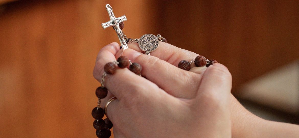 Due mani tengono un rosario di legno. Si vede la croce del rosario.