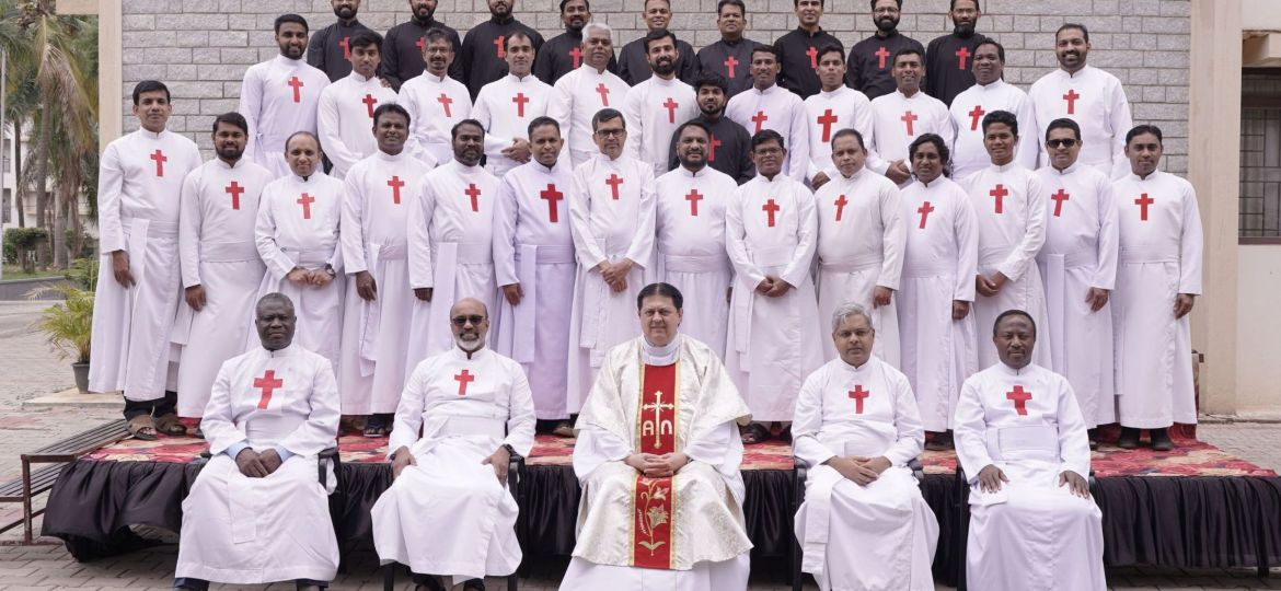 Gruppo di uomini vestiti con abiti sacerdotali bianchi con la croce rossa di san Camillo. Gli uomini sono disposti frontalmente su un palco di 4 file. Sullo sfondo c'è un muro di mattoni grigi. Nella fila più in basso ci sono solo cinque uomini e sono seduti. Tutti gli altri sono in piedi. L'uomo al centro degli uomini seduti indossa gli abiti per la messa. Al posto della croce rossa ha una lunga striscia rossa sull'abito bianco.