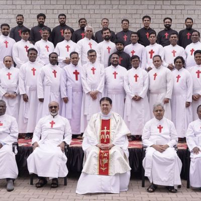Gruppo di uomini vestiti con abiti sacerdotali bianchi con la croce rossa di san Camillo. Gli uomini sono disposti frontalmente su un palco di 4 file. Sullo sfondo c'è un muro di mattoni grigi. Nella fila più in basso ci sono solo cinque uomini e sono seduti. Tutti gli altri sono in piedi. L'uomo al centro degli uomini seduti indossa gli abiti per la messa. Al posto della croce rossa ha una lunga striscia rossa sull'abito bianco.
