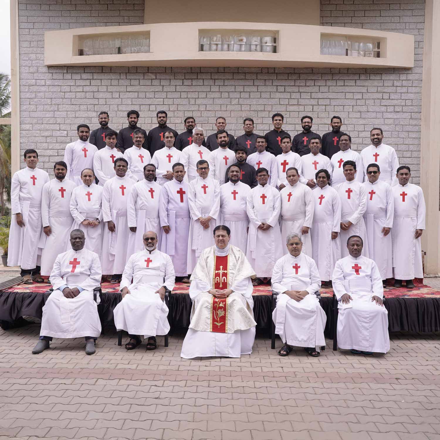 Gruppo di uomini vestiti con abiti sacerdotali bianchi con la croce rossa di san Camillo. Gli uomini sono disposti frontalmente su un palco di 4 file. Sullo sfondo c'è un muro di mattoni grigi. Nella fila più in basso ci sono solo cinque uomini e sono seduti. Tutti gli altri sono in piedi. L'uomo al centro degli uomini seduti indossa gli abiti per la messa. Al posto della croce rossa ha una lunga striscia rossa sull'abito bianco.