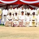 Un gruppo di sacerdoti camilliani con gli abiti bianchi e gialli da cerimonia posano su un prato arido per una foto. Dietro di loro si vede un tendone rosso.