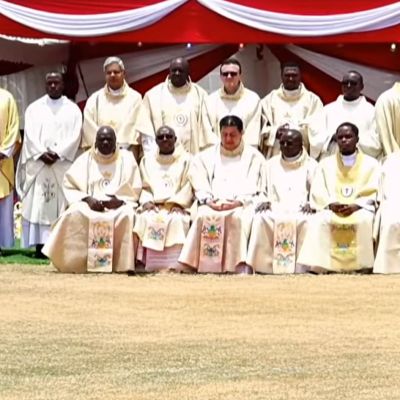 Un gruppo di sacerdoti camilliani con gli abiti bianchi e gialli da cerimonia posano su un prato arido per una foto. Dietro di loro si vede un tendone rosso.