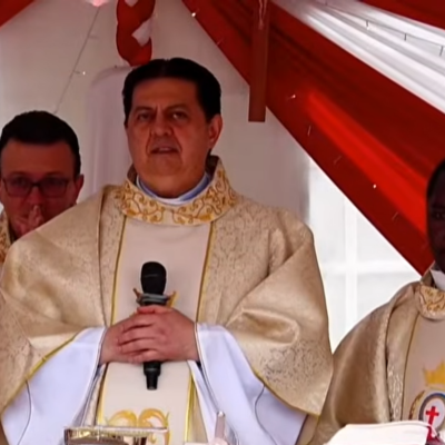 Tre sacerdoti stanno celebrando la messa. Sono vestiti con abiti bianchi e dorati. Si trovano sotto a un tendone bianco e rosso