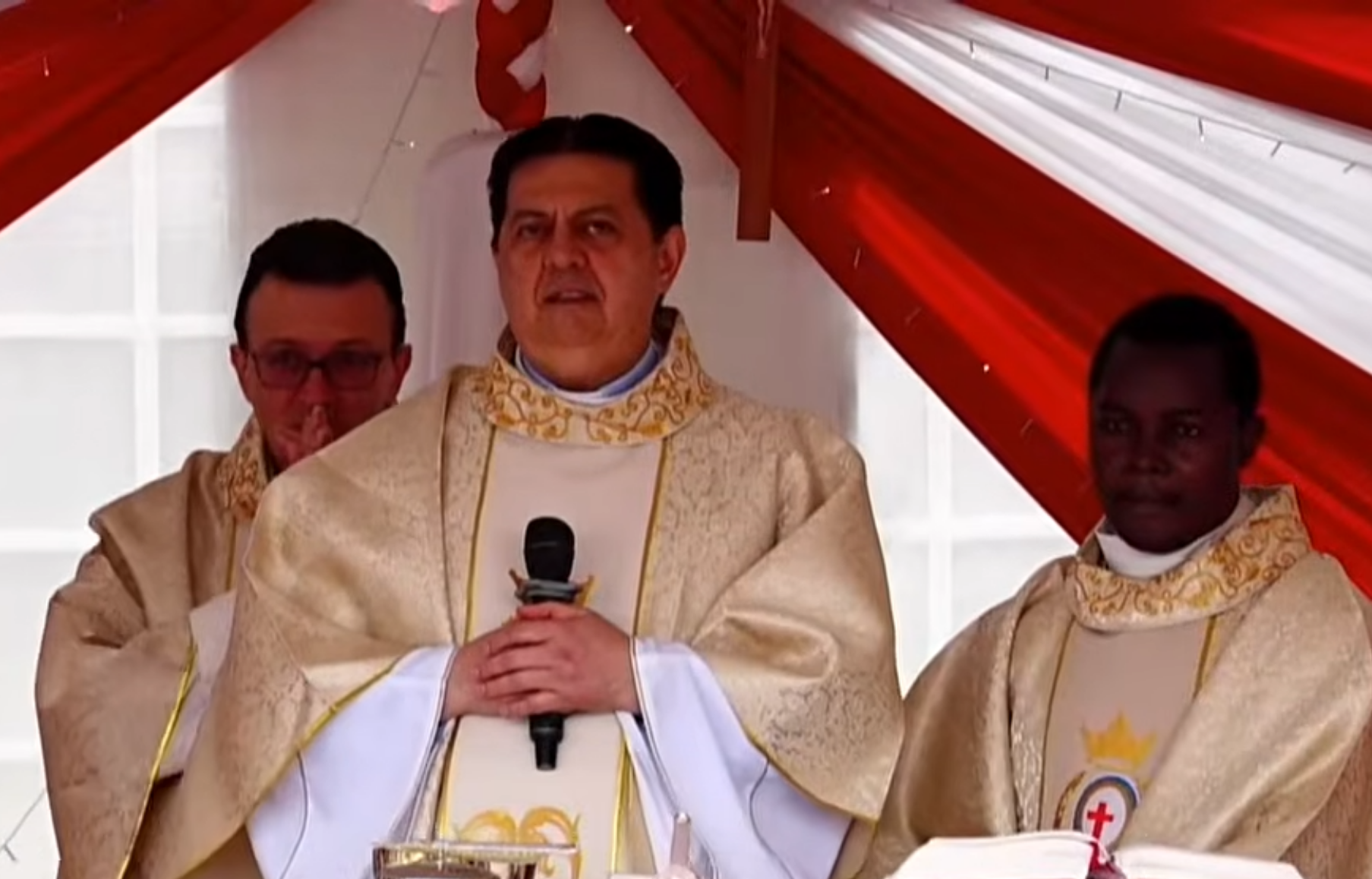 Tre sacerdoti con la tunica bianca celebrano la messa sotto un tendone rosso e bianco