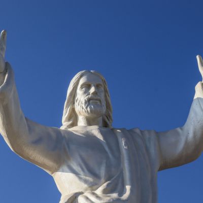 Una statua bianca di Cristo ha le mani alzate. La statua si trova all'aperto. Intorno si vede solo il cielo azzurro quasi viola
