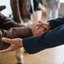 Nella foto si vedono duo uomini che si stringono le mani in modo informale. Uno dei due uominio ha una giacca marrone, l'altro ha una giacca blu. Sononuno di fronte all'altro. Quello con la giacca blu sostiene il gomito dell'uomo con la giacca marrone.