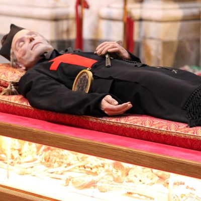 San Camillo vestito dcon la tunica nera e la croce rossa sul petto è sdraiato su un materasso rosso. Sotto di lui, illuminate in giallo, si vedono delle ossa.