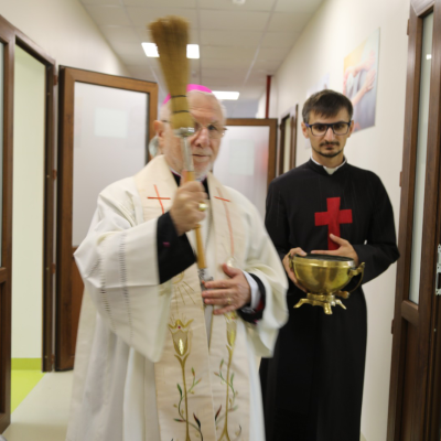 Un vescovo vestito di bianco con zucchetto viola benedice un locale. Accanto a lui c'è un religioso camilliano vestito con la tonaca nera con la croce rossa.