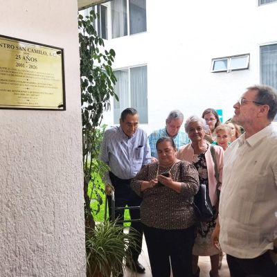 Delle persone guardano una targa dorata sul muro. In primo piano di profilo c'è un uomo con i capelli grigi, gli occhiali e una camicia bianca.