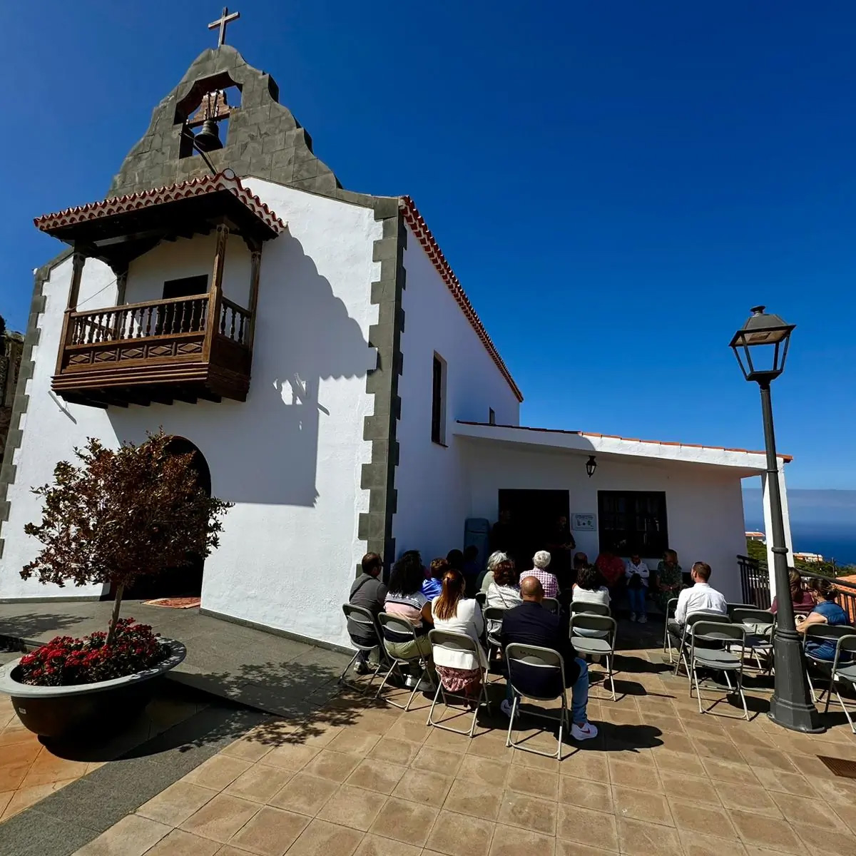 Accanto a una piccola chiesa bianca, un gruppo di persone ascolta seduta all'aperto un relatore. Il cielo è blu. Il terreno è marrone